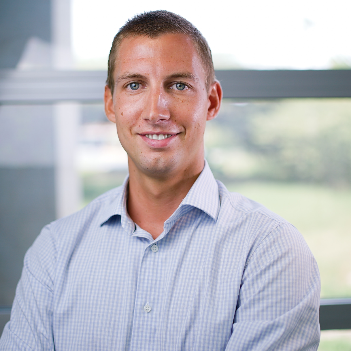 A smiling man in a checkered shirt poses for a photo in a bright environment with large windows.