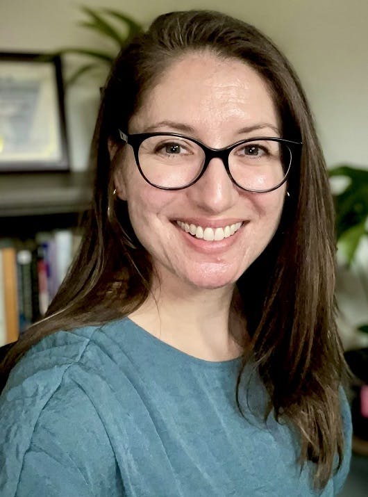 A person with glasses smiling, wearing a blue shirt, in an indoor setting with books and a plant in the background.