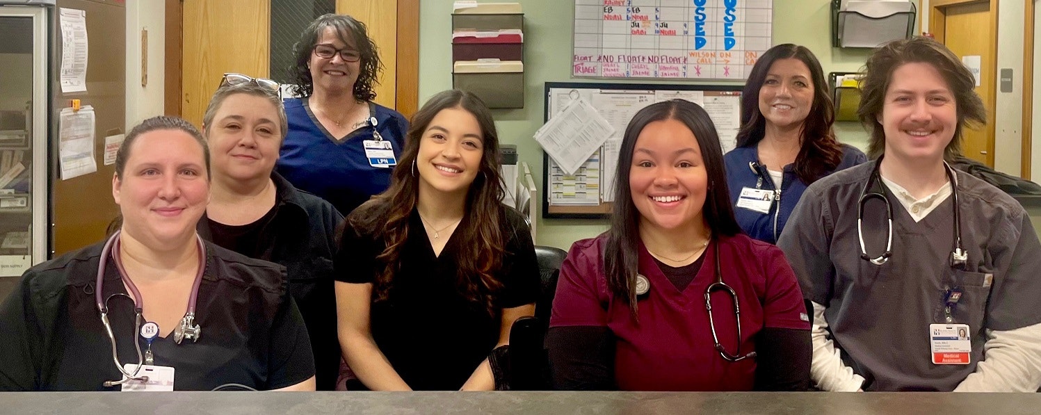 A group of seven smiling healthcare workers in uniforms with stethoscopes and name badges, standing in an office setting.