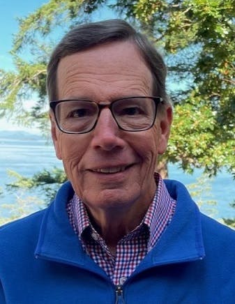 A person wearing glasses and a blue jacket, standing outdoors with a tree and water in the background.