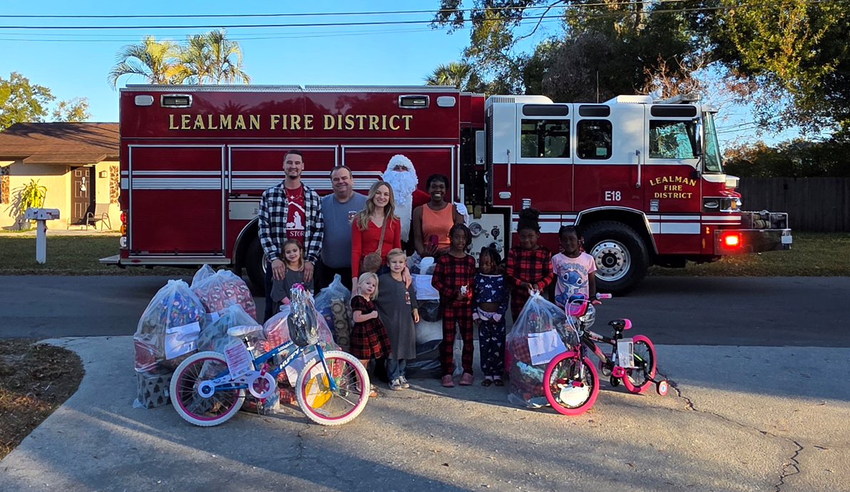 A fire truck is parked with a group of people and children, celebrating the holiday season with gifts and bikes.