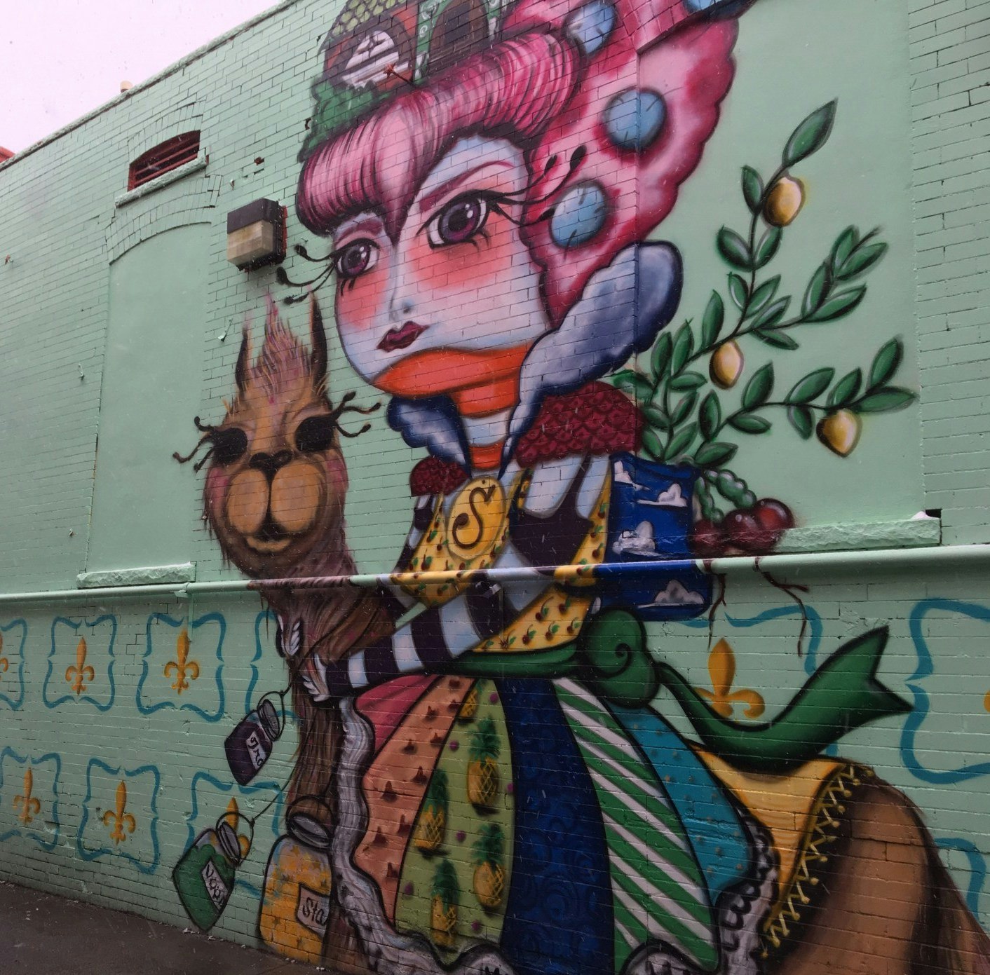 A colorful mural featuring a whimsical character with pink hair, holding a llama, with a green leafy background on a brick wall.