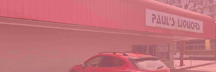 A red car is parked outside a brick building with a sign reading "Paul's Liquors."