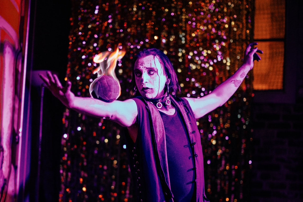A performer with theatrical makeup holds a flaming object, set against a sparkly background.