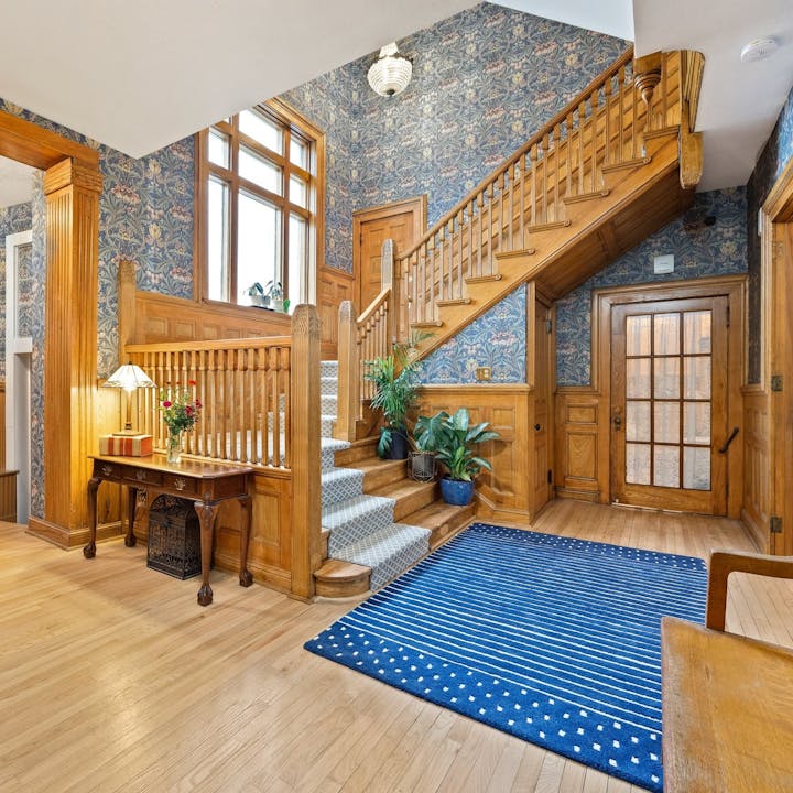 Wooden staircase with patterned wallpaper, plants, a blue rug, and a wooden table with a lamp and flowers in an elegant room.