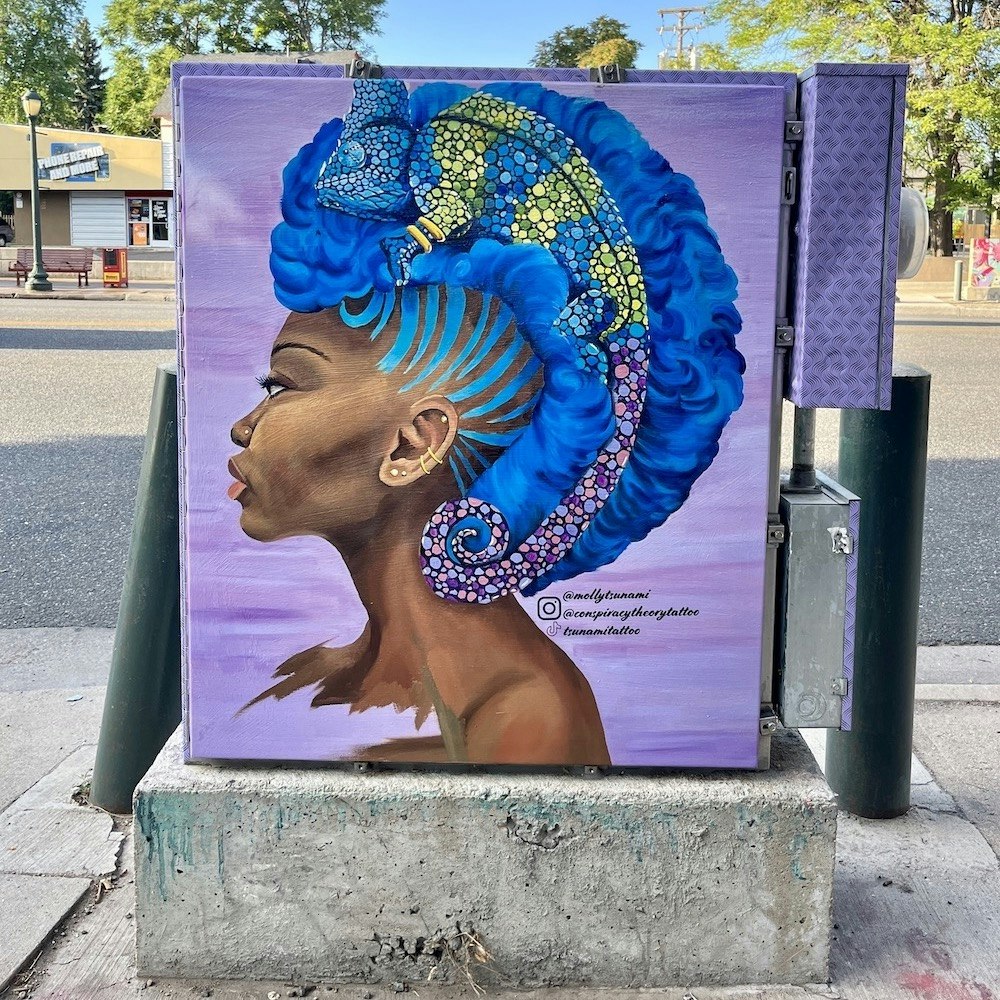 Street art of a side profile portrait with bright blue hair adorned with patterns and a chameleon, painted on an urban utility box.