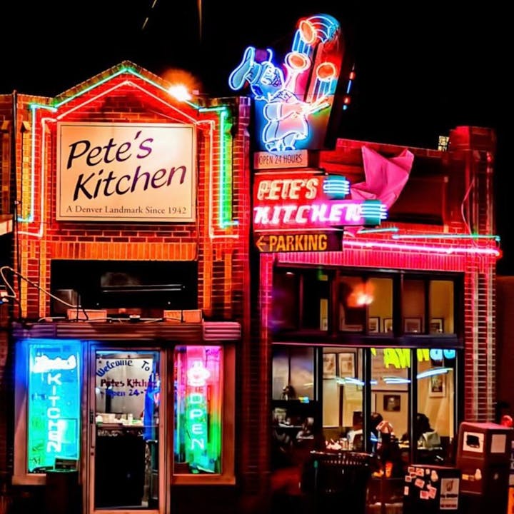 Colorful neon-lit exterior of "Pete's Kitchen" diner, open 24 hours, with signs highlighting its status as a Denver landmark since 1942.