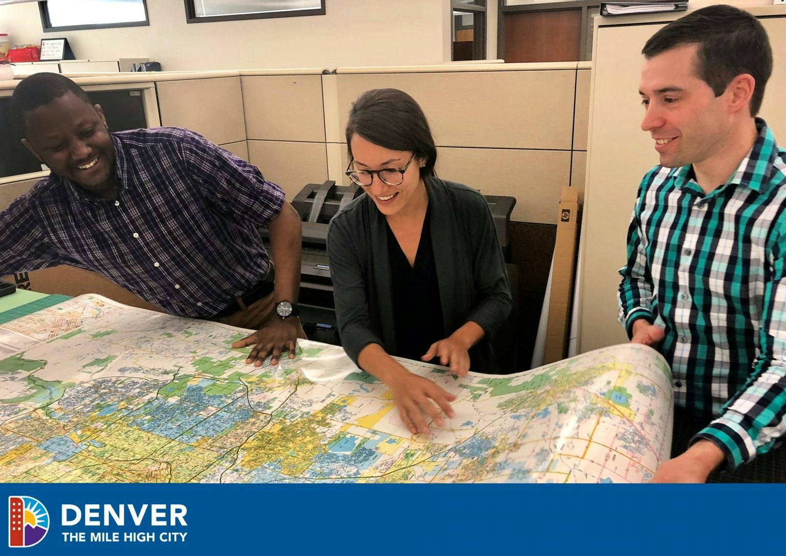 Three people smiling and looking at a large map in an office setting.