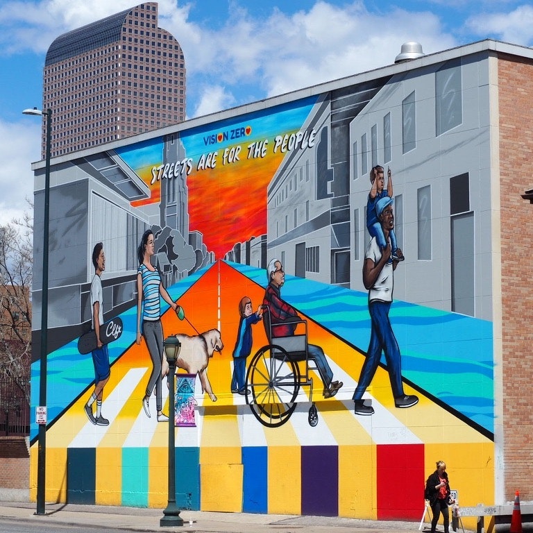 Mural reading "STREETS ARE FOR THE PEOPLE" shows diverse group, including people with a dog, on a colorful street scene.