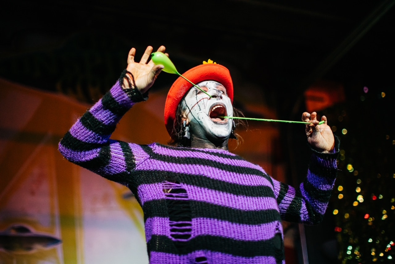 A person with face paint and a red hat stretches a green balloon, wearing a striped sweater on stage.