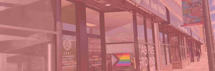 A storefront with signage for "Above Ground" and "Gool," featuring a Progress Pride flag and colorful street banners. People are walking by.