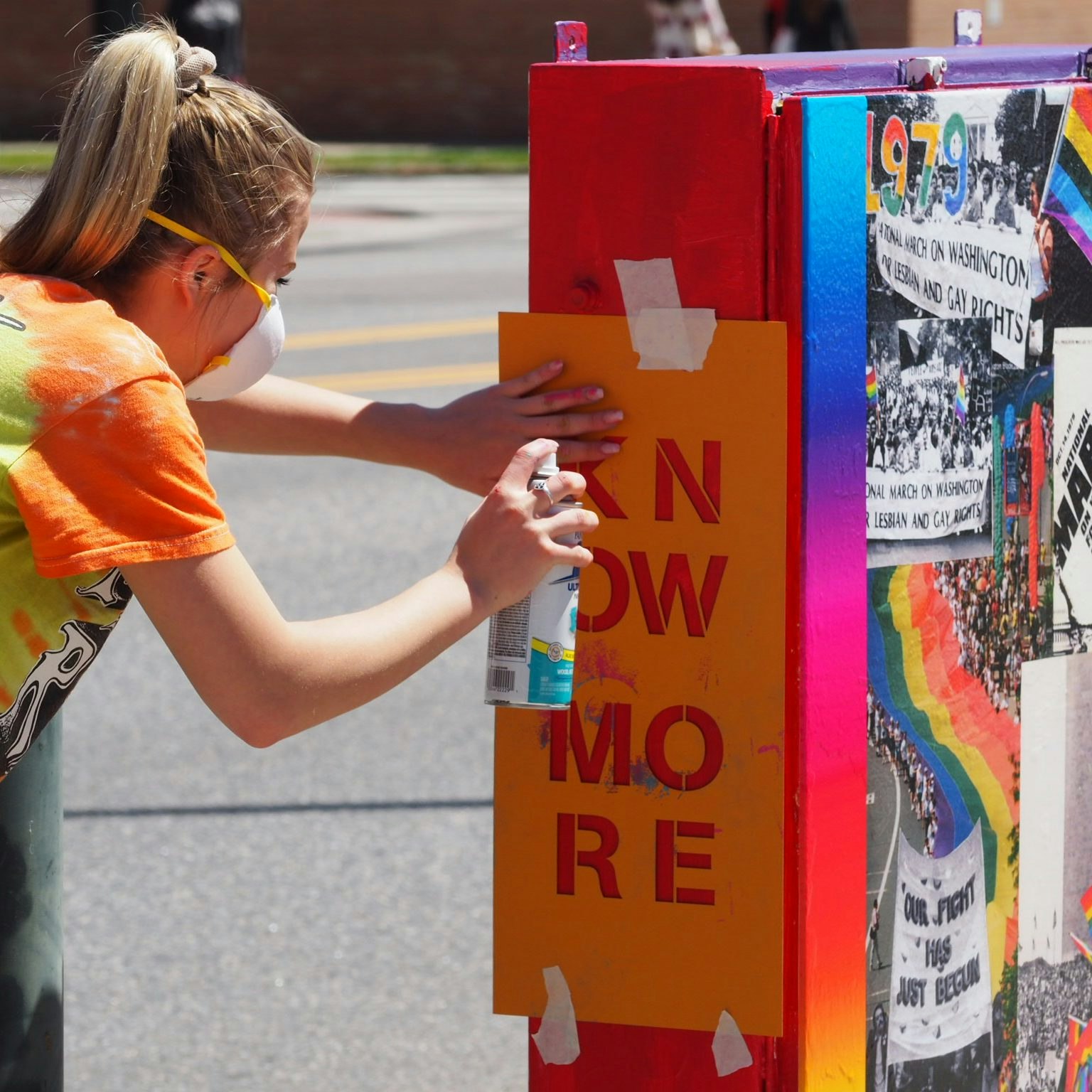 A person spray-paints "KNOW MORE" using a stencil on a colorful box with images and rainbow colors.