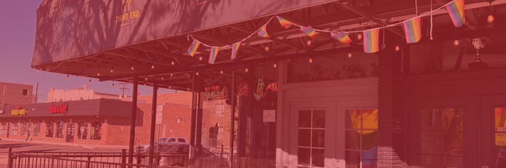 Outdoor cafe with rainbow flags, neon signs, and string lights, next to a street with signs visible.