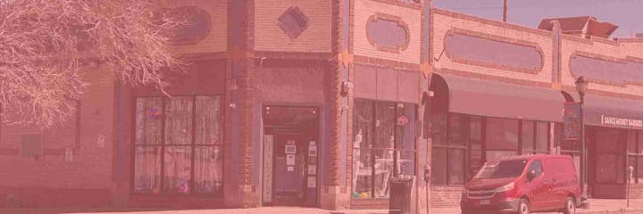 A brick building on a corner with storefronts, a tree, and a red van parked in front.