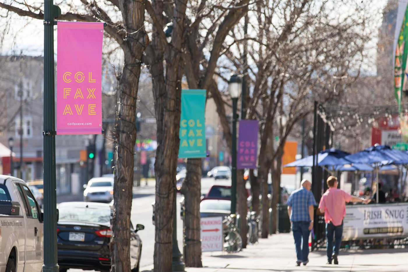 Street view of Colfax Ave with colorful banners, cars parked, and people walking by a restaurant with blue umbrellas.