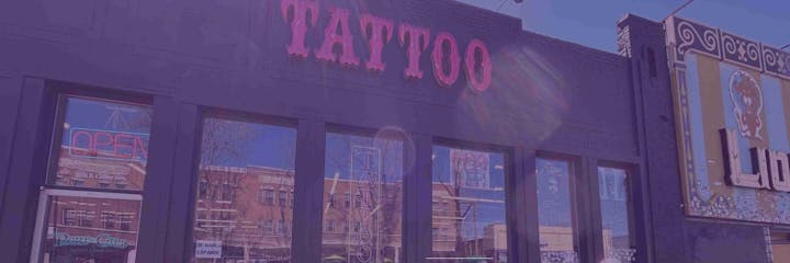 Storefront with "Tattoo" sign, neon "Open" sign, window posters, and ornate decoration on adjacent building.