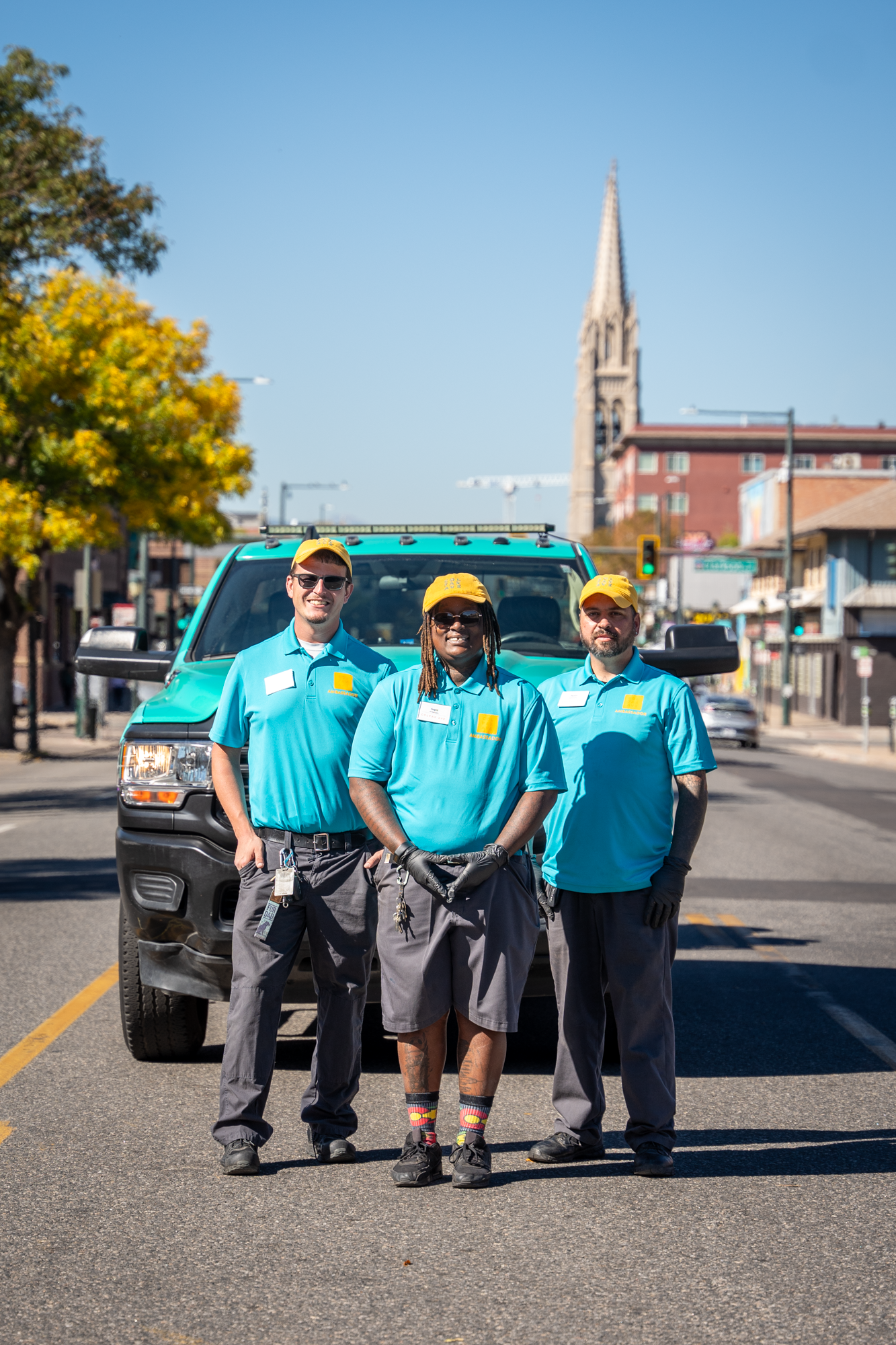 Three people in matching uniforms and caps stand in front of a parked vehicle on a sunny street.