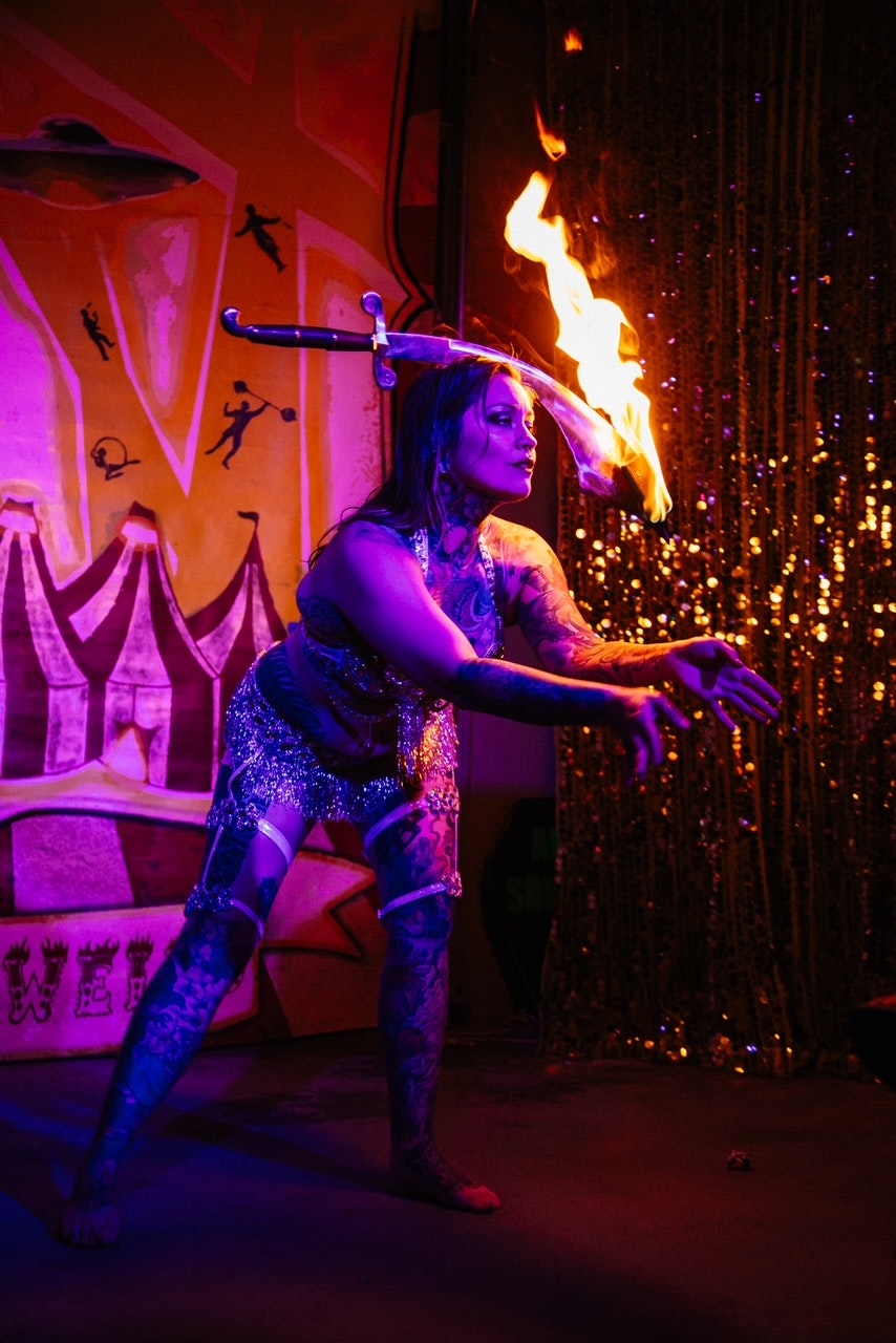 Performer in costume balances a flaming sword on their head, colorful backdrop with circus and UFO art.