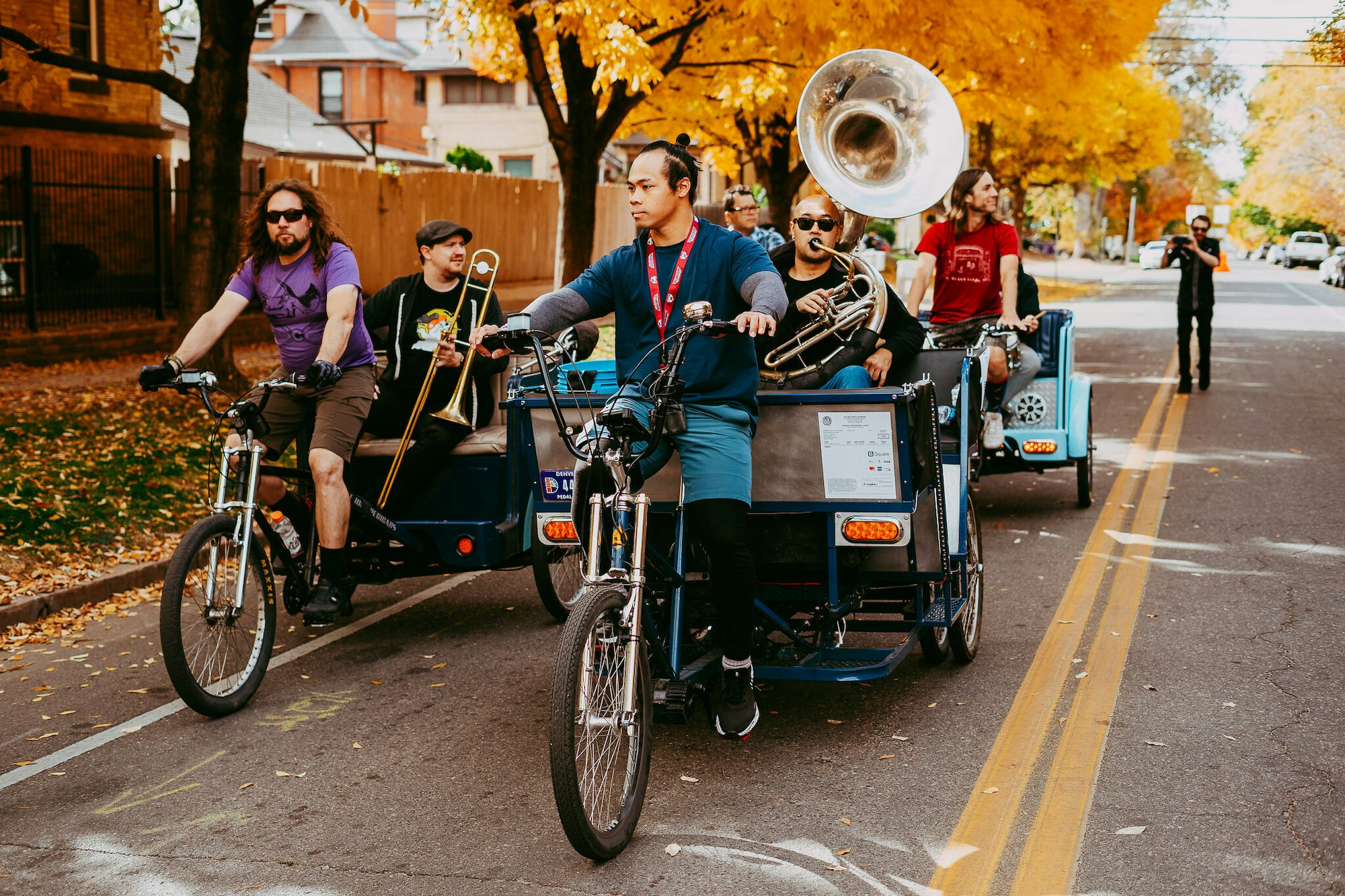 A group of people riding bicycles with musical instruments, including a trombone and a sousaphone, on a tree-lined street in autumn.