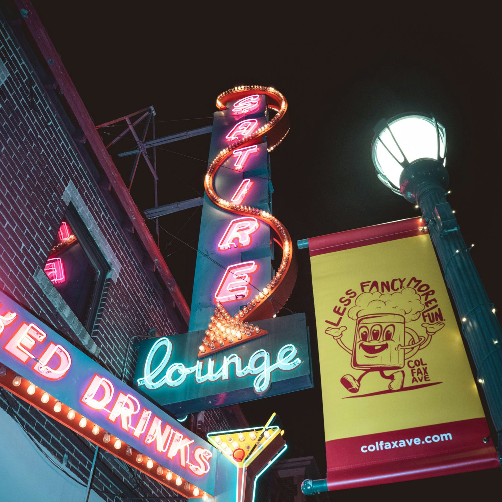 Neon signs for "Satire Lounge" and a drink, with a banner reading "Less Fancy More Fun" and a cartoon character on Colfax Ave at night.