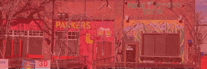 Red brick building with murals, signs for a bar, and banners. Trees and colorful wall art with text visible.