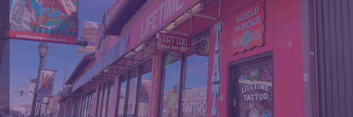 Street view of a tattoo shop with colorful signage, banners, and art on Colfax Avenue.