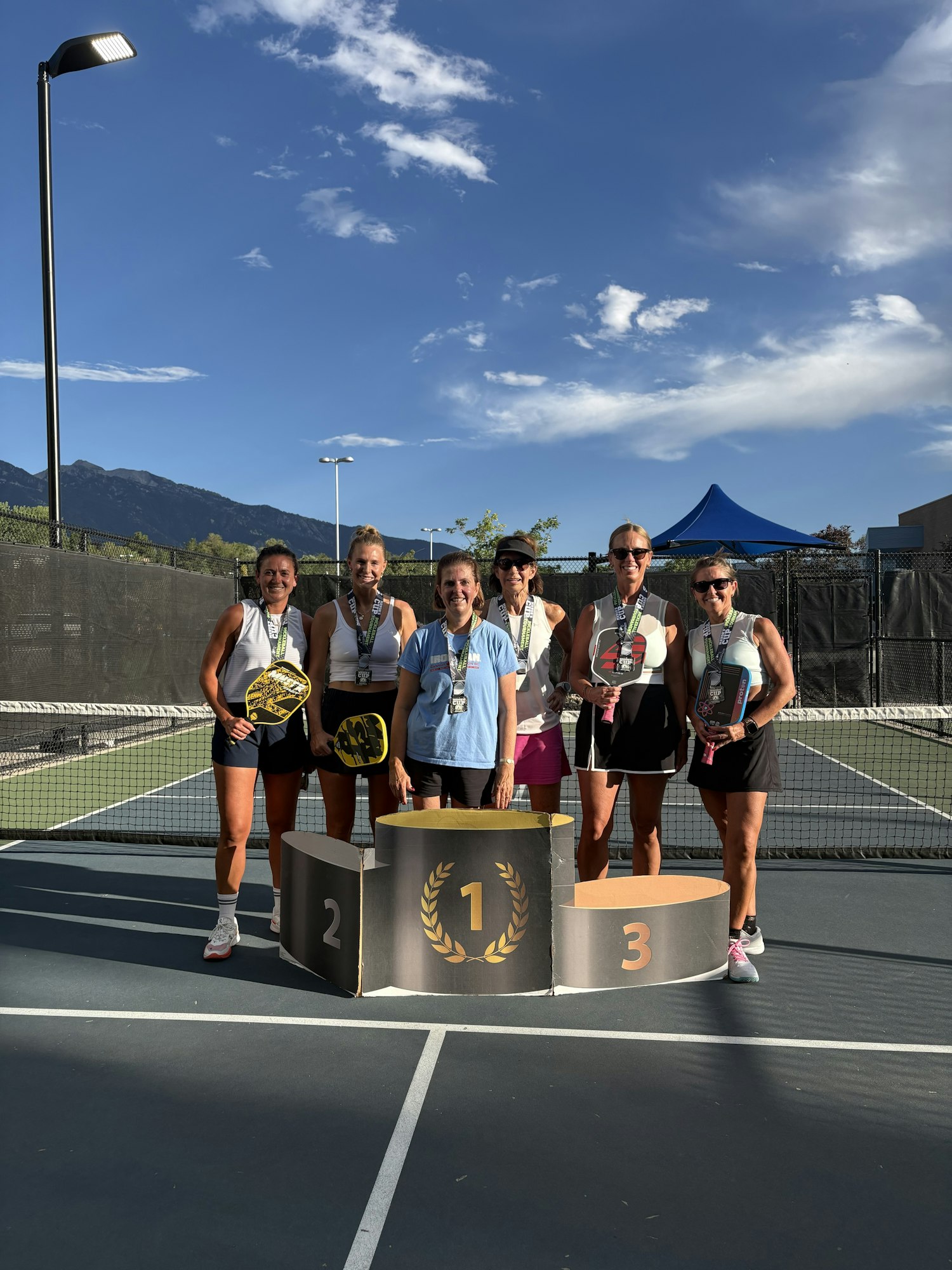 A group of six women stand on a tennis court by a podium, celebrating a pickleball tournament with medals and paddles.