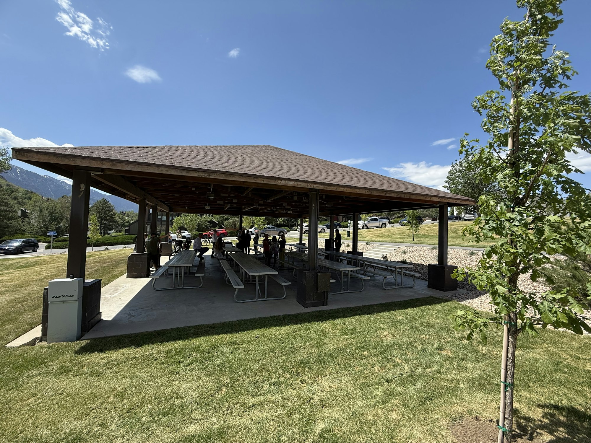 A pavilion with picnic tables, people gathered, green lawn, and a blue sky, set in a park-like area.