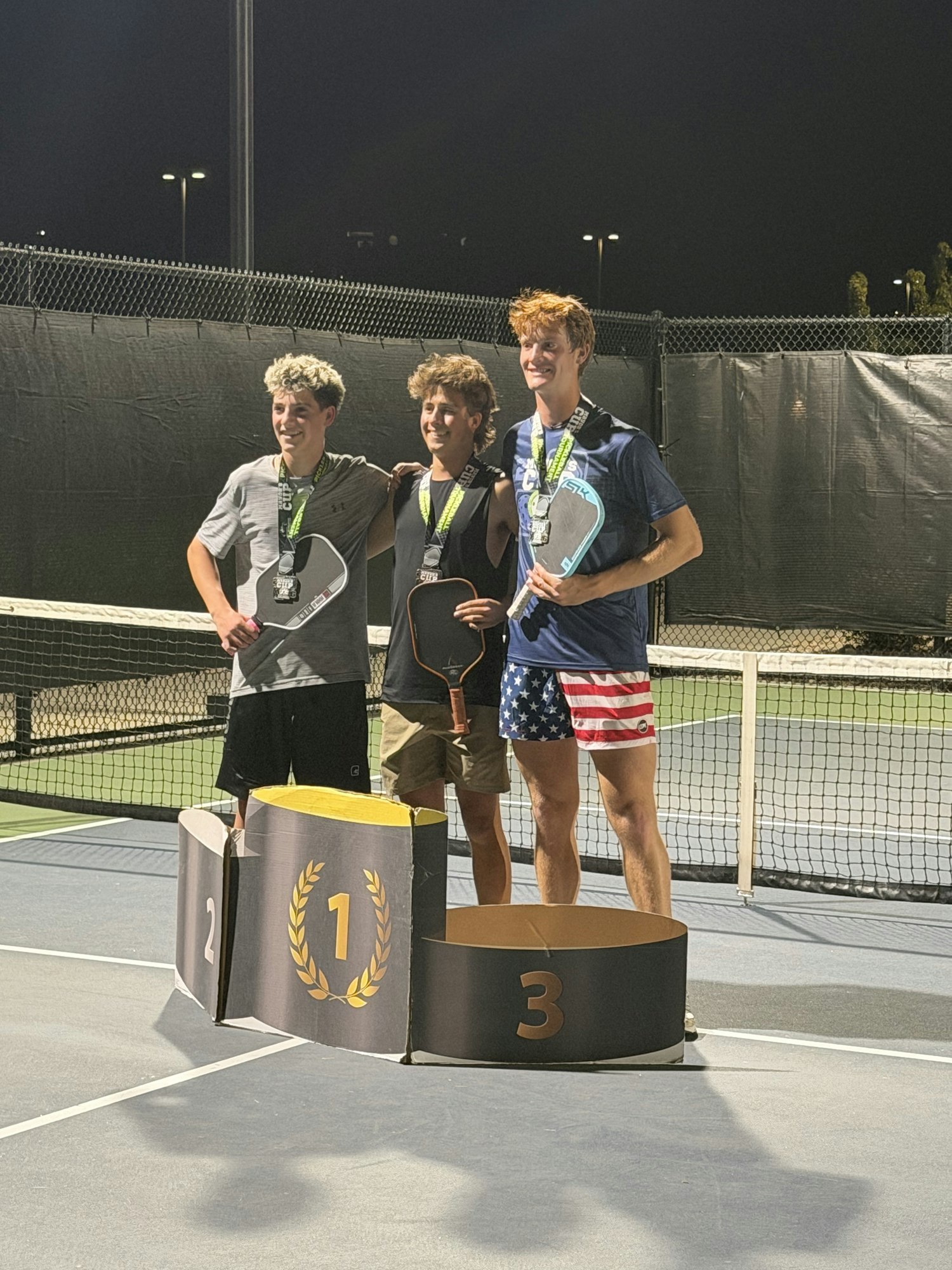 Three young men stand on a podium at a tennis court, smiling while holding paddles and wearing medals. It's night time.