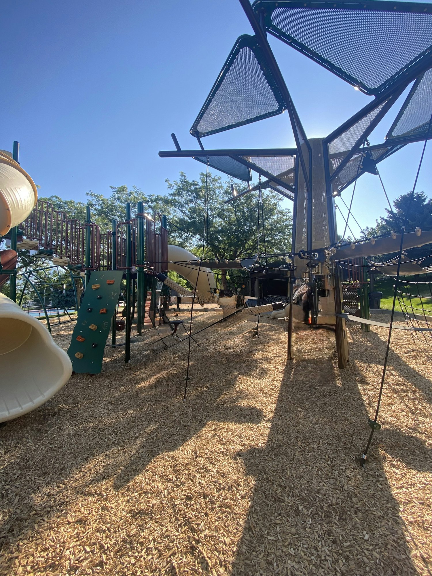Playground with climbing structures, slides, and nets on woodchip ground. Sunny day with clear sky and trees in the background.