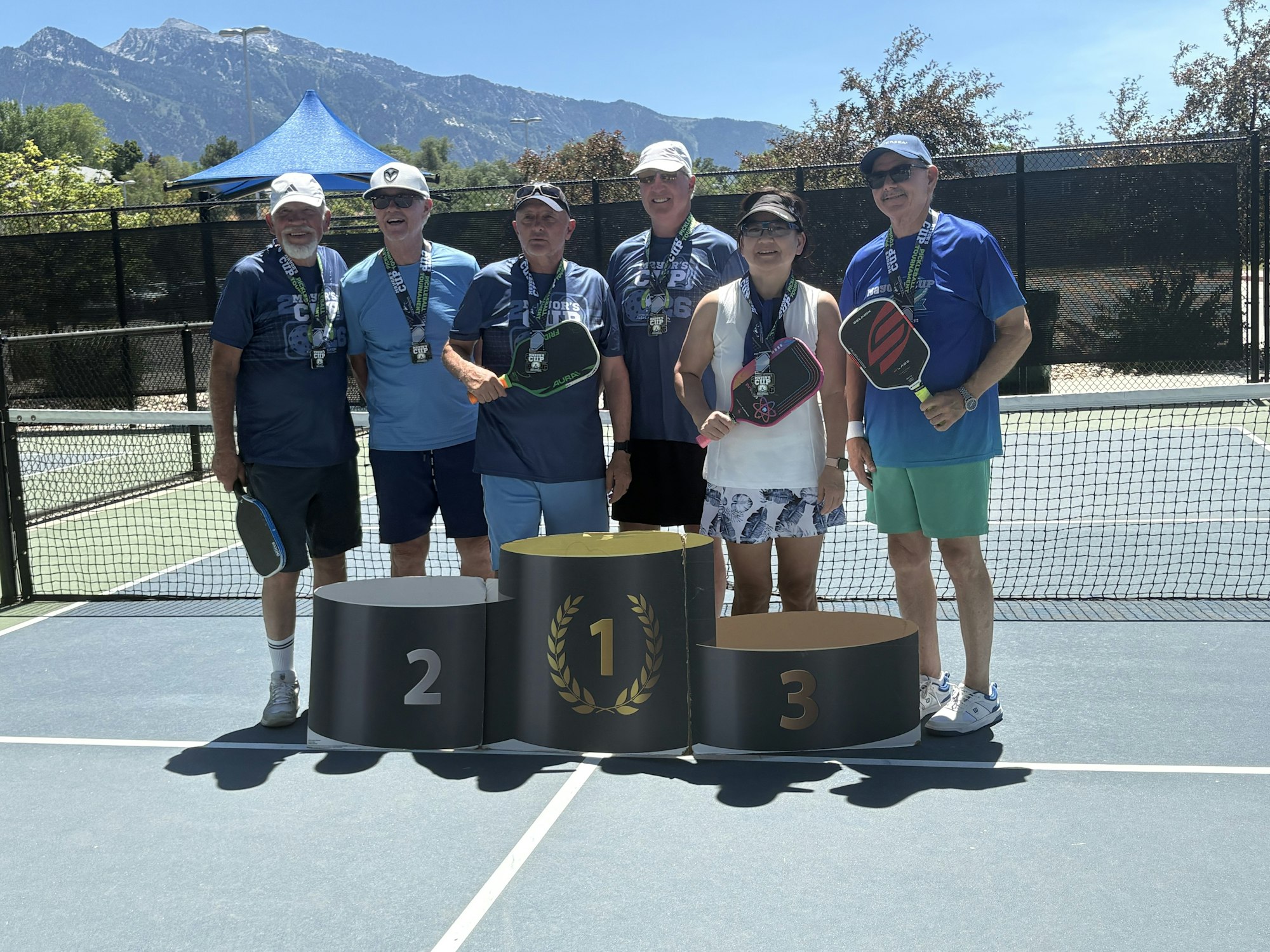 A group of six people stands on a tennis court, posing with paddles and medals near podiums labeled 1st, 2nd, and 3rd.