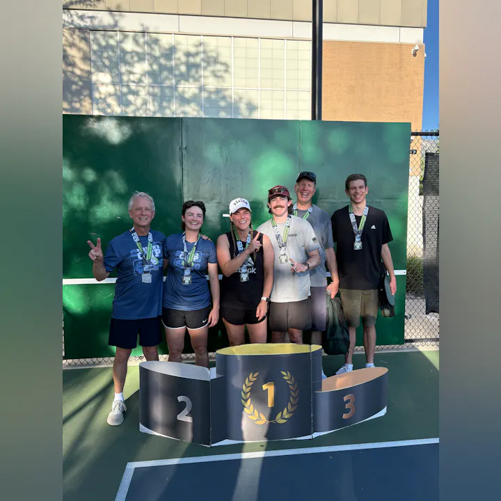 A group of six people poses with medals on a podium after a competition, celebrating a victory outdoors.