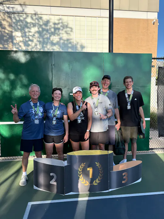 A group of six people poses with medals on a podium after a competition, celebrating a victory outdoors.