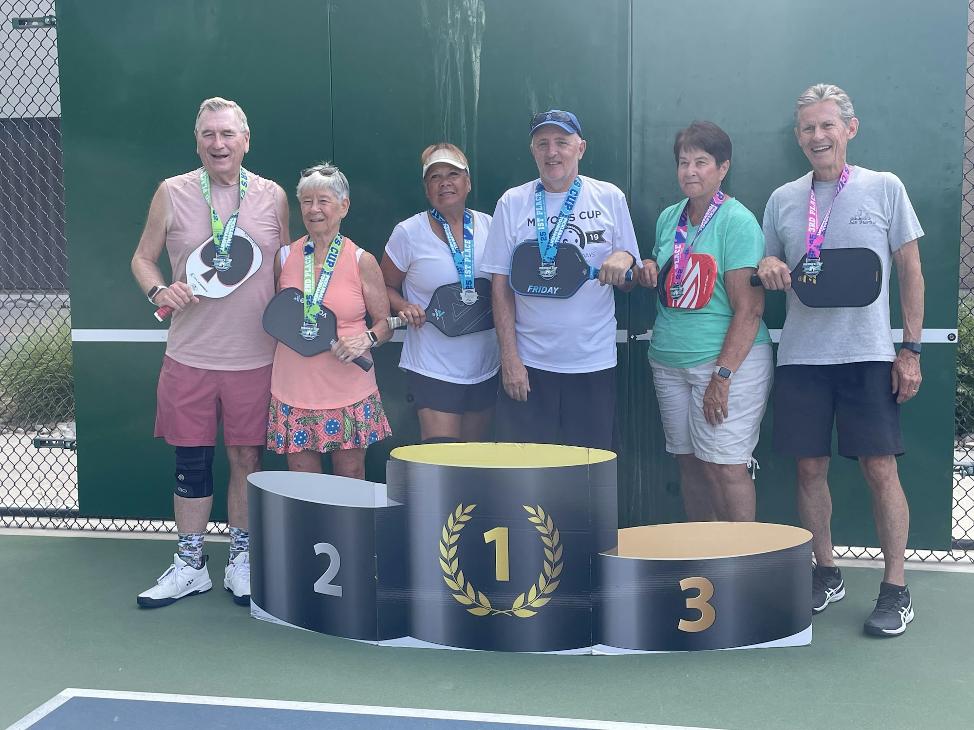 Six people with paddles and medals, smiling on a pickleball podium outdoors.