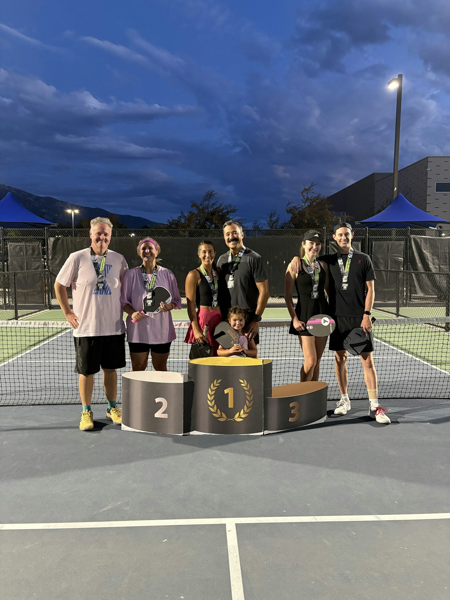A group of people poses with medals and a trophy on a podium at a tennis or pickleball event, under a twilight sky.