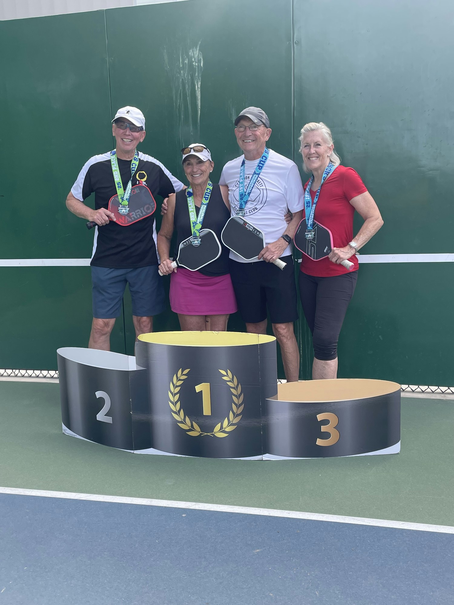 Five people posing with pickleball paddles and medals on a podium with positions 1, 2, and 3.