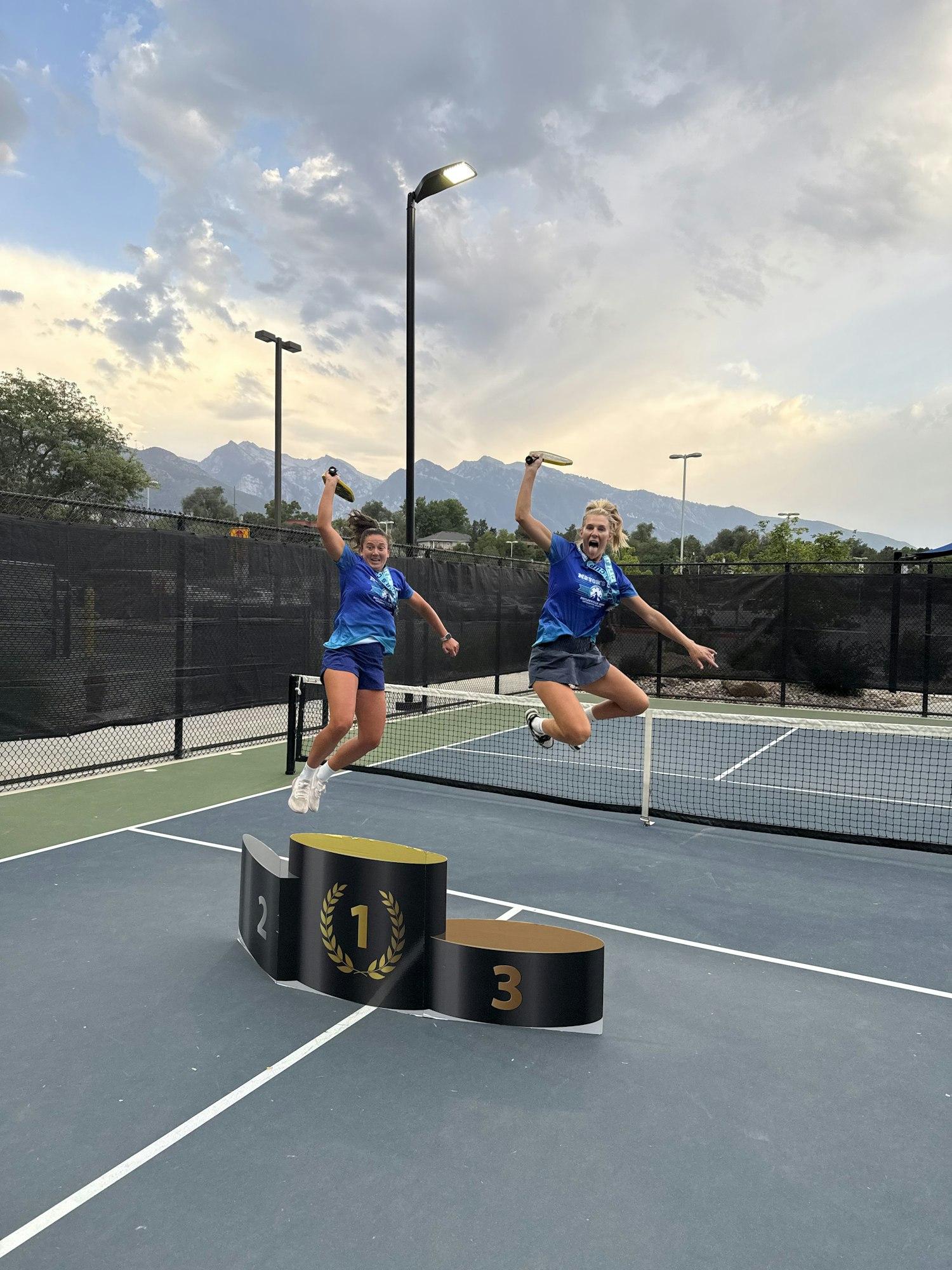 Two people jumping joyfully on a tennis court next to a podium with their paddles.