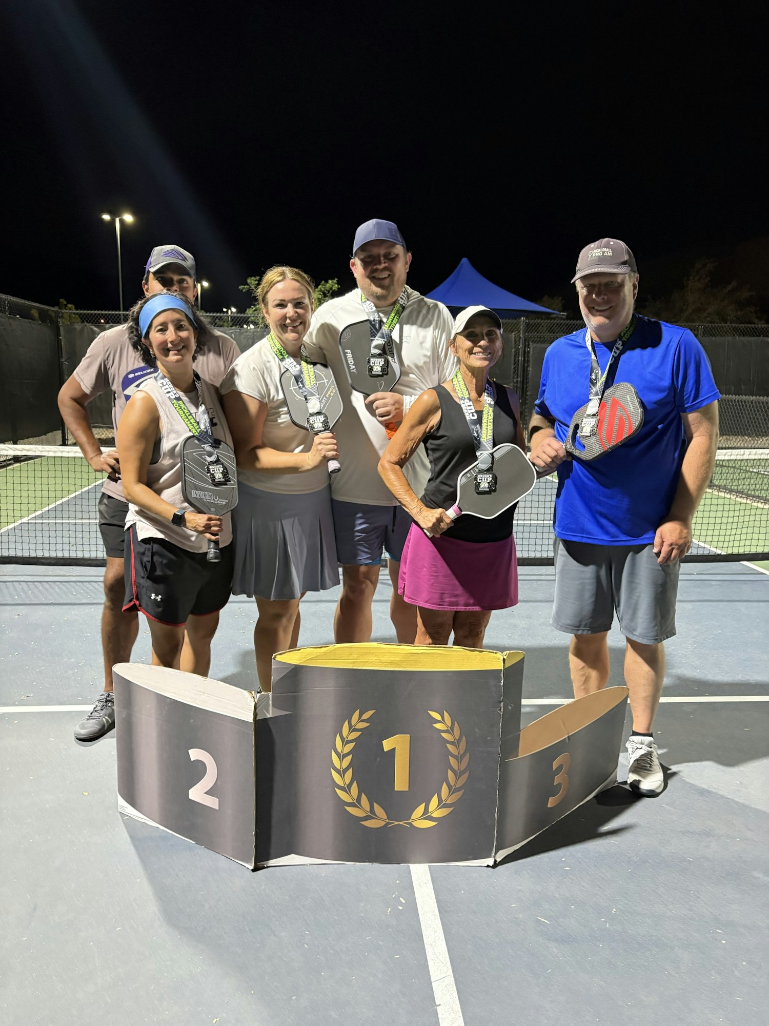 A group of six people poses at night with medals and paddles near a podium, celebrating a pickleball event.