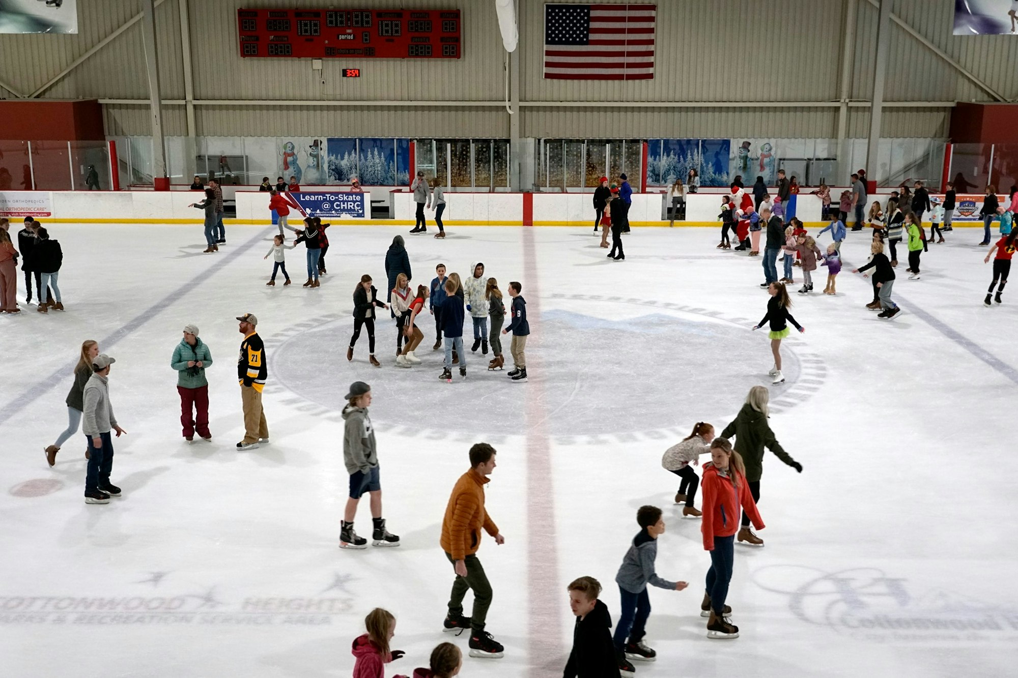 An ice skating rink filled with people of all ages enjoying skating, with a large flag and festive decorations in the background.