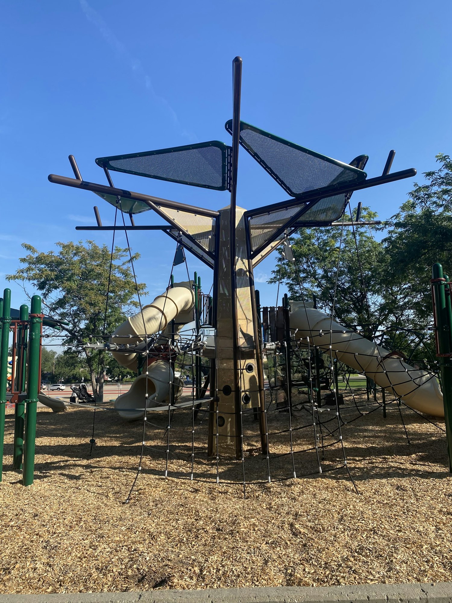 Playground structure with climbing nets, slides, and shade panels under a clear sky.