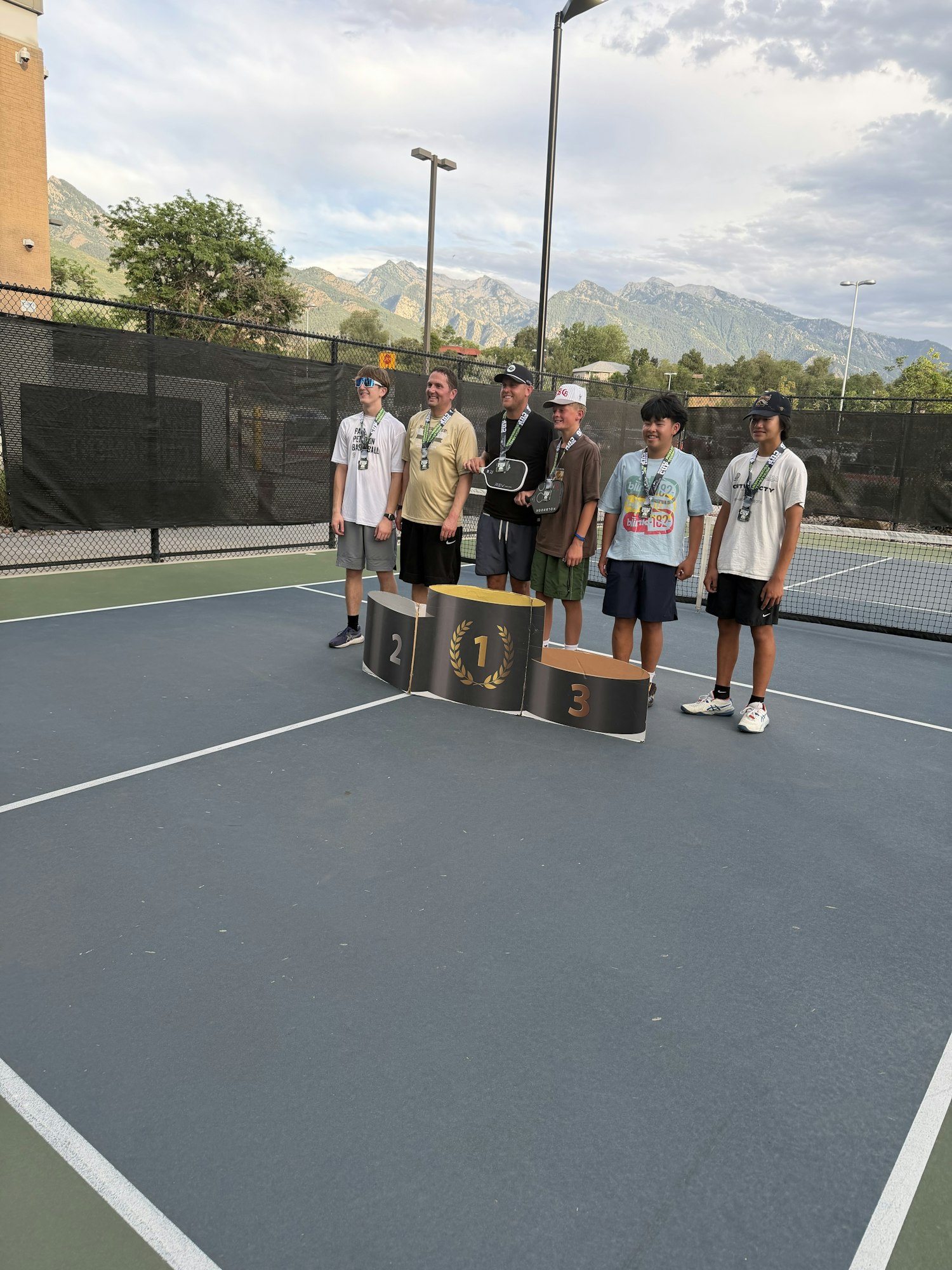A group of six people stands on a tennis court by a podium, celebrating after a tournament, with mountains in the background.