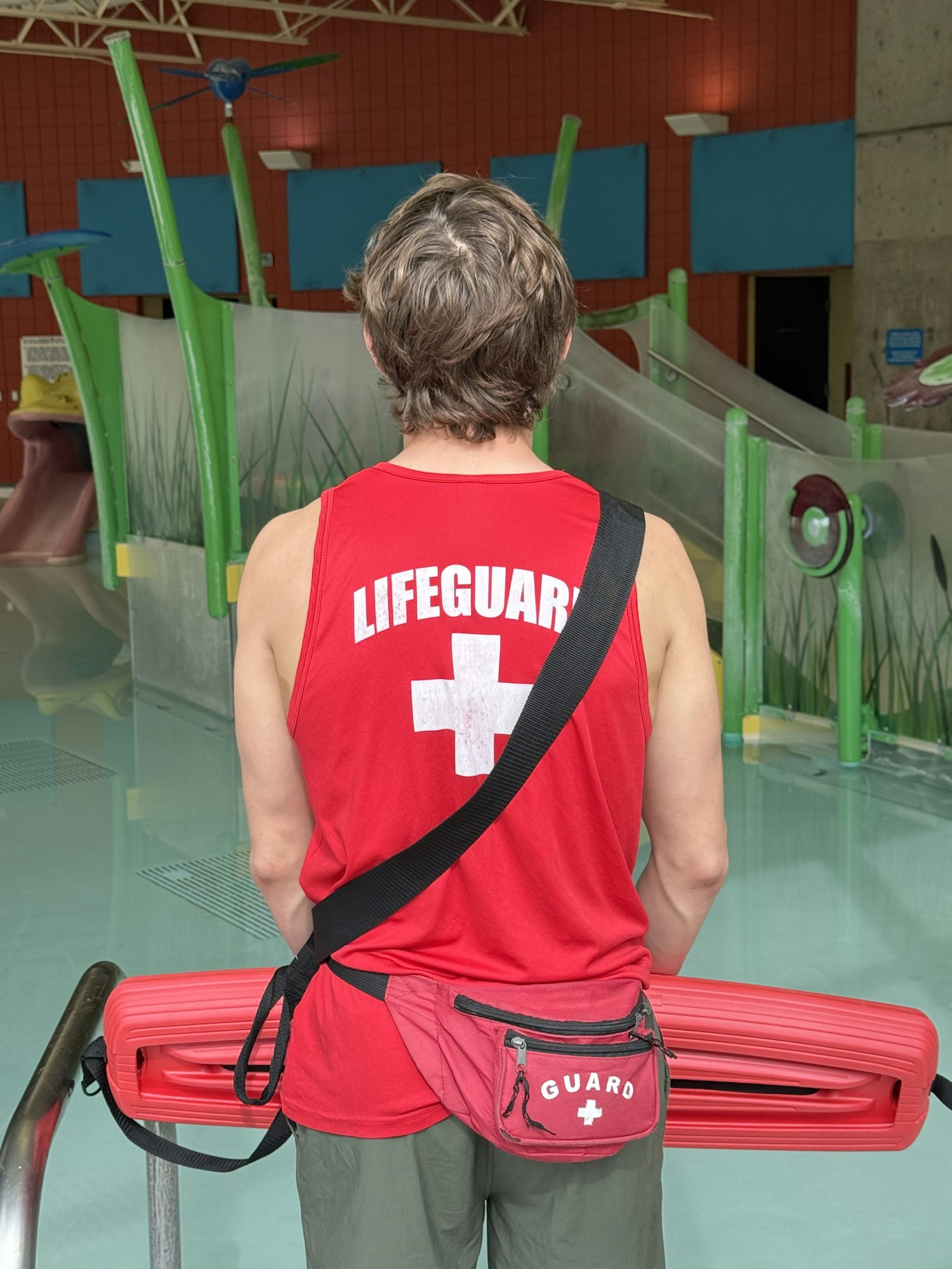 A lifeguard in a red tank top stands near a pool area, holding a rescue flotation device. Colorful water play equipment is in the background.
