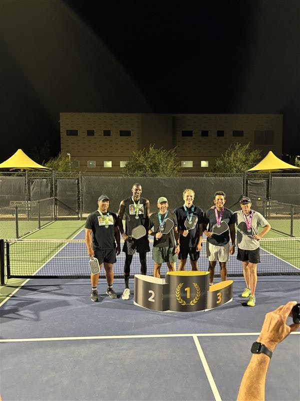 A group of six people with medals and paddles, standing on a podium at a pickleball court at night.