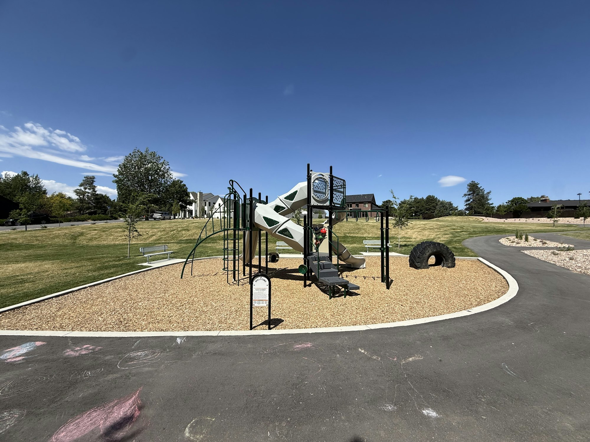 The image shows a playground with a slide, climbing structure, and a tire, set in a grassy park under a clear blue sky.