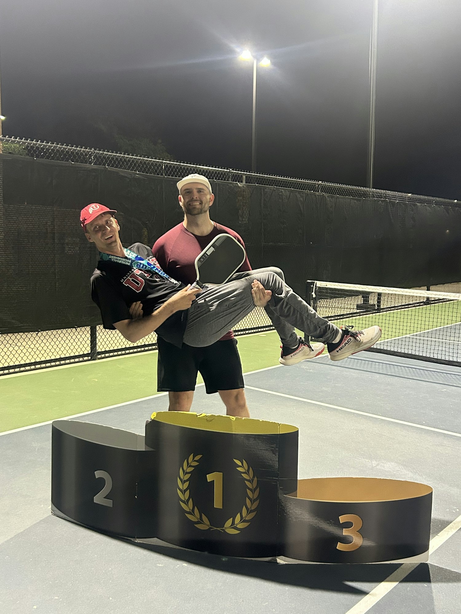 Two people on a tennis court, one carrying the other by a podium marked 1, 2, 3.