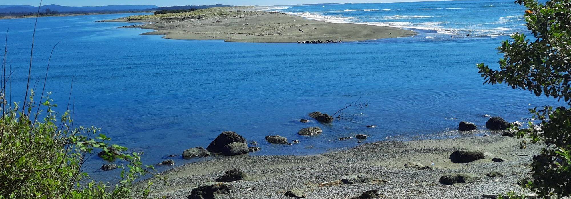 A serene landscape with a blue river, sandy shore, scattered rocks, and greenery, framed by distant hills and ocean waves.