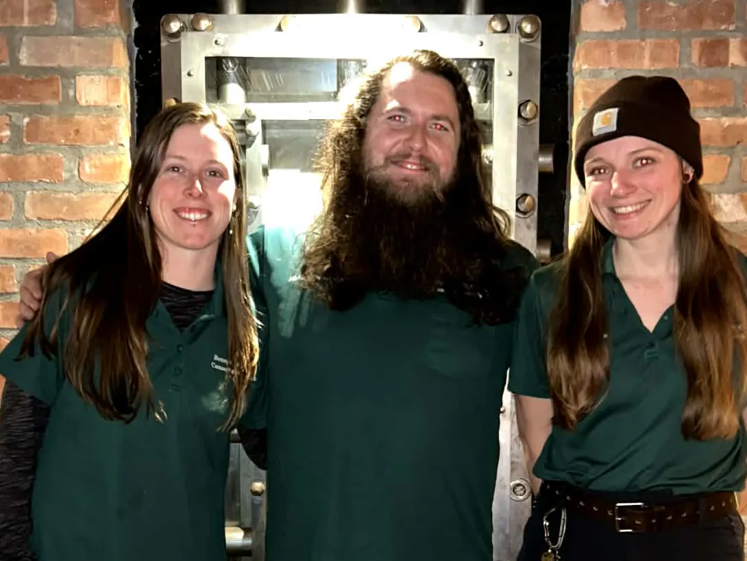 Three individuals in green shirts pose together in front of a vintage-style vault door, smiling at the camera.
