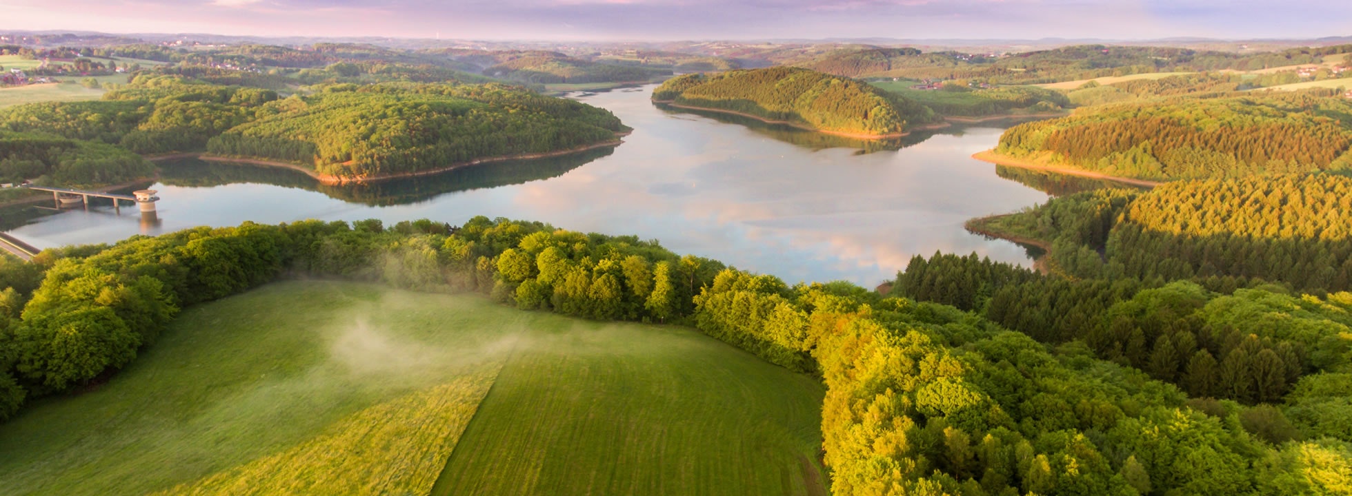 Scenic aerial view of a lake surrounded by lush green forests and rolling hills, under a cloud-filled sky.