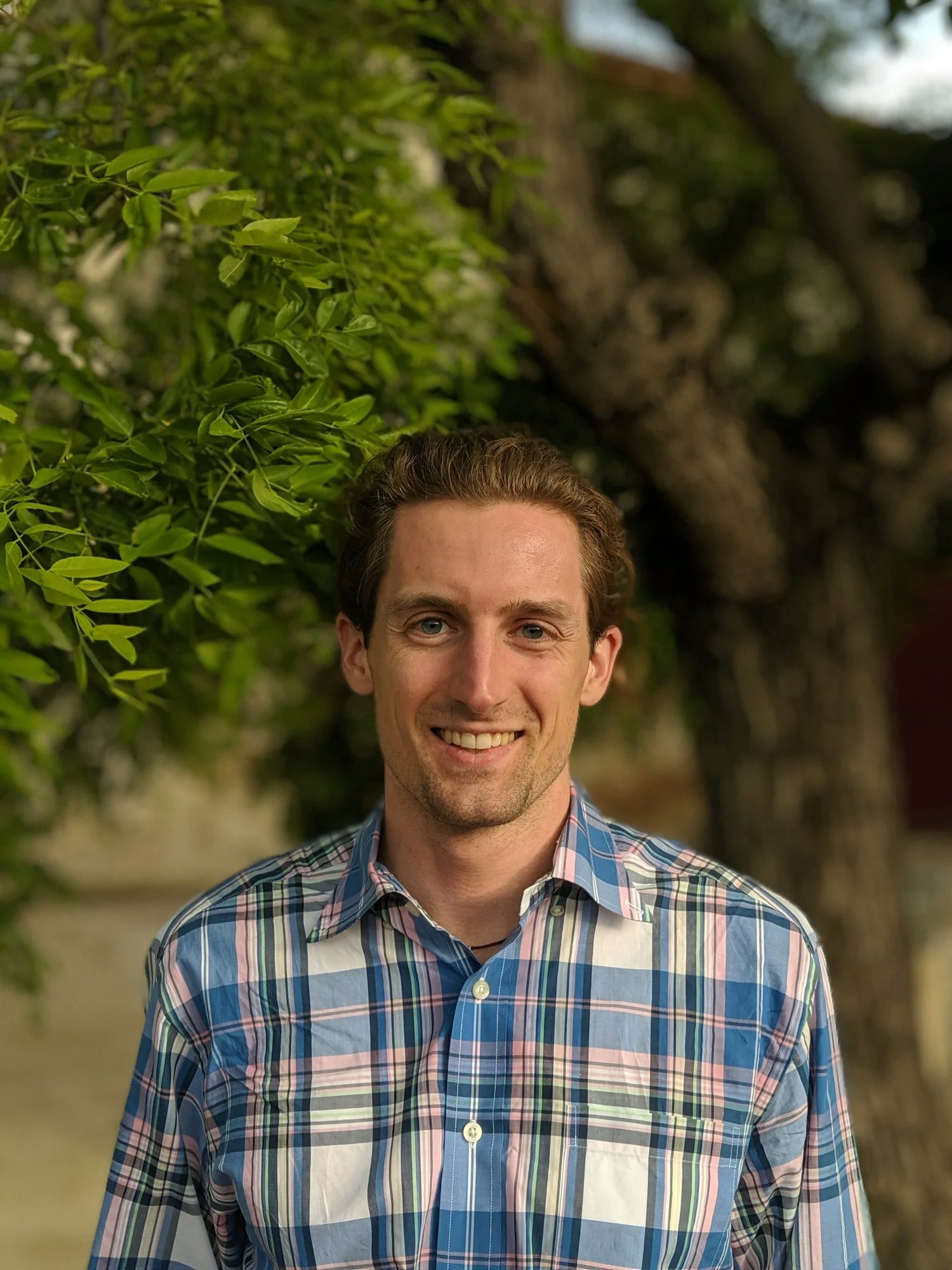 A person in a plaid shirt smiles in front of green leaves and a tree.