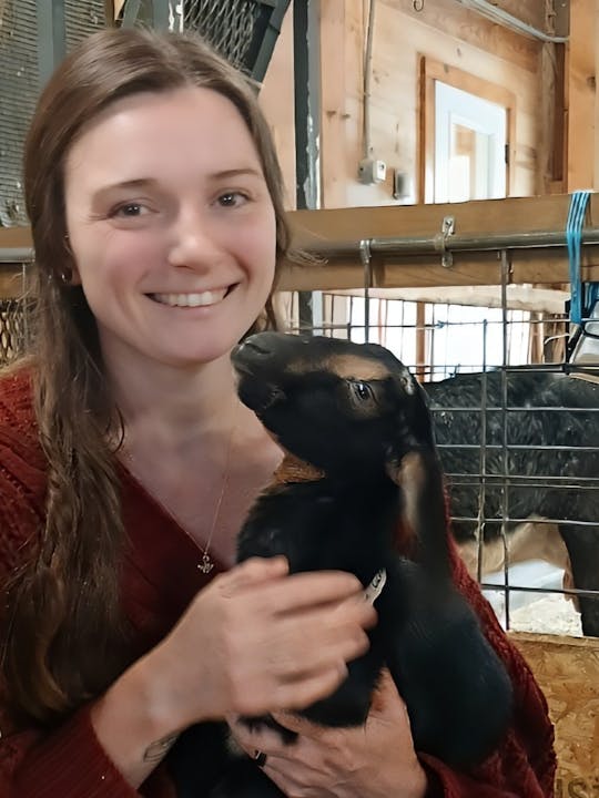 A person is smiling while holding a baby goat inside a barn.